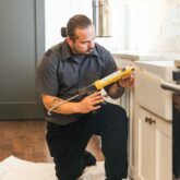 Handyman applying silicone onto a farmhouse sink in a white kitchen.