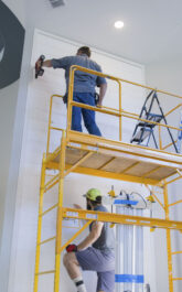 Two Handyman standing on scaffolding installing a custom wood façade to a fireplace